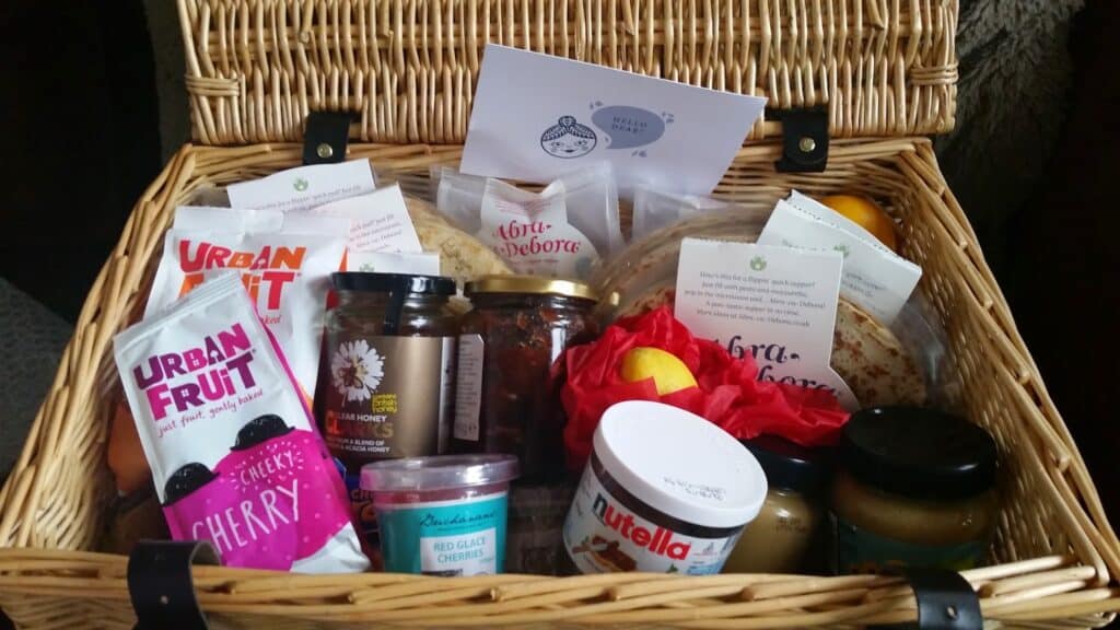 A wicker hamper filled with various food items, including packets of Urban Fruit snacks, jars of honey, Nutella, preserves, flatbreads, a wrapped lemon, biscuits, and a card on top. The contents are neatly arranged and look colourful and inviting.