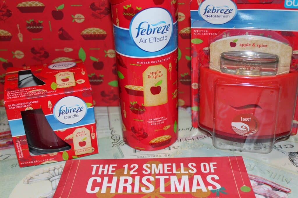 A display of Febreze apple & spice scented products, including a candle, air spray, Set & Refresh, and a jar candle. The items are red with apple graphics. Behind them is apple-themed wrapping paper. A sign in front reads "THE 12 SMELLS OF CHRISTMAS.