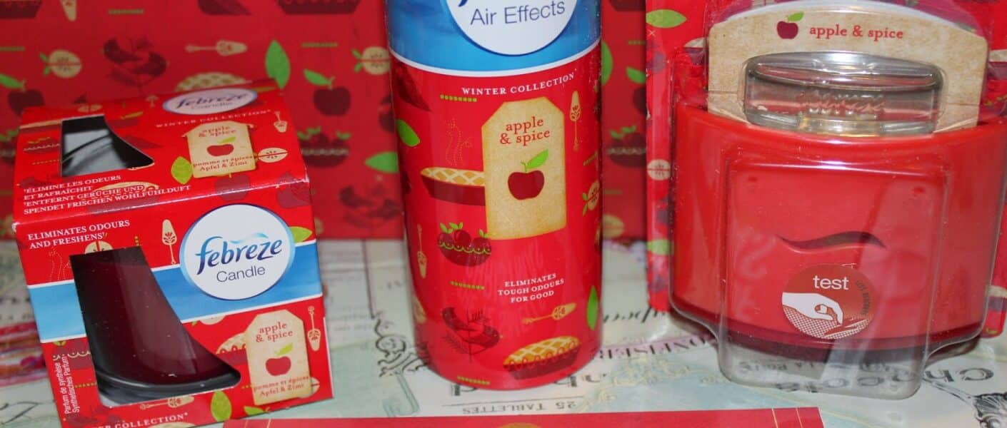 A display of Febreze apple & spice scented products, including a candle, air spray, Set & Refresh, and a jar candle. The items are red with apple graphics. Behind them is apple-themed wrapping paper. A sign in front reads "THE 12 SMELLS OF CHRISTMAS.
