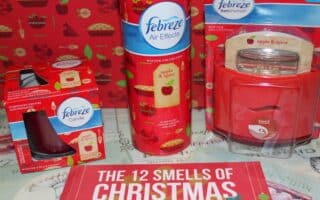 A display of Febreze apple & spice scented products, including a candle, air spray, Set & Refresh, and a jar candle. The items are red with apple graphics. Behind them is apple-themed wrapping paper. A sign in front reads "THE 12 SMELLS OF CHRISTMAS.