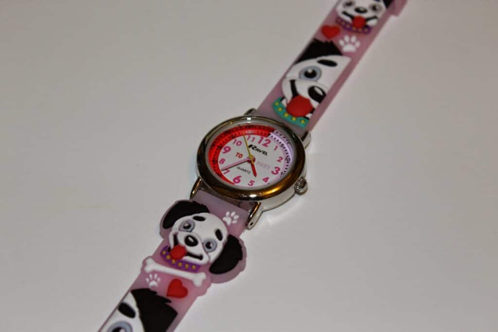 A children’s wristwatch with a pink strap decorated with cartoon Dalmatian puppies, paw prints, and red hearts. The round watch face shows both standard and 24-hour time, with black numbers, a red inner ring, and black hour and minute hands.