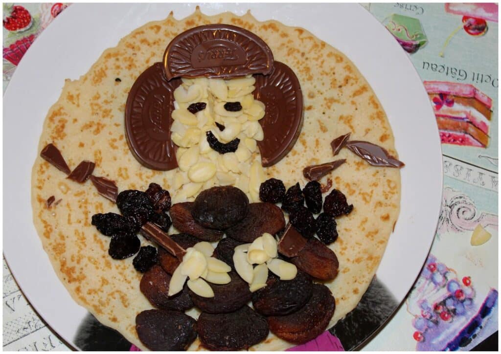 A pancake on a white plate is decorated to resemble a face and body. Chocolate pieces form hair and a hat; flaked almonds, sultanas, and dried apricots create eyes, a mouth, arms, and a dress; all sit on a colourful, patterned table mat.