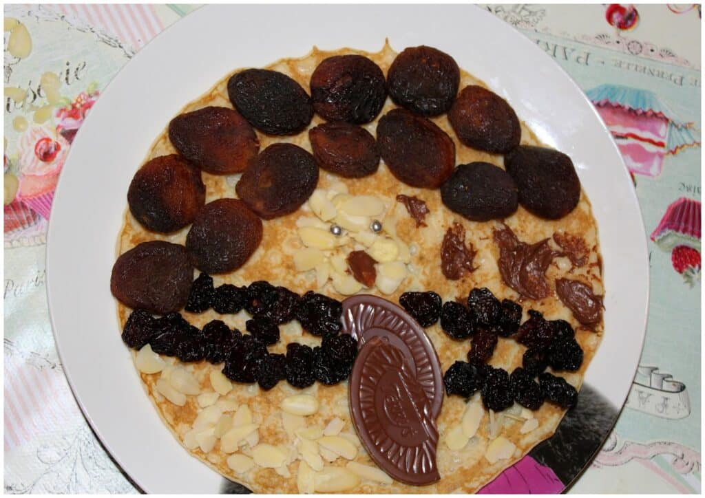 A pancake on a white plate is decorated to resemble a face, using dried apricots for hair, raisins for a mouth, slivered almonds for facial features, chocolate spread for eyes, and a chocolate segment for a smiling mouth. The background shows a colourful tablecloth.