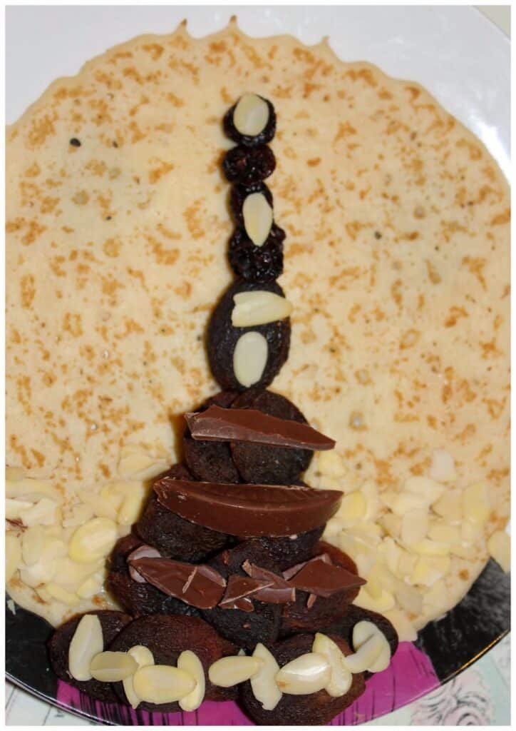 A round crêpe on a plate is decorated with pieces of chocolate, almonds, and raisins arranged upright to resemble a minaret or tower, with scattered sliced almonds at the base and a pink serviette visible in the bottom right corner.