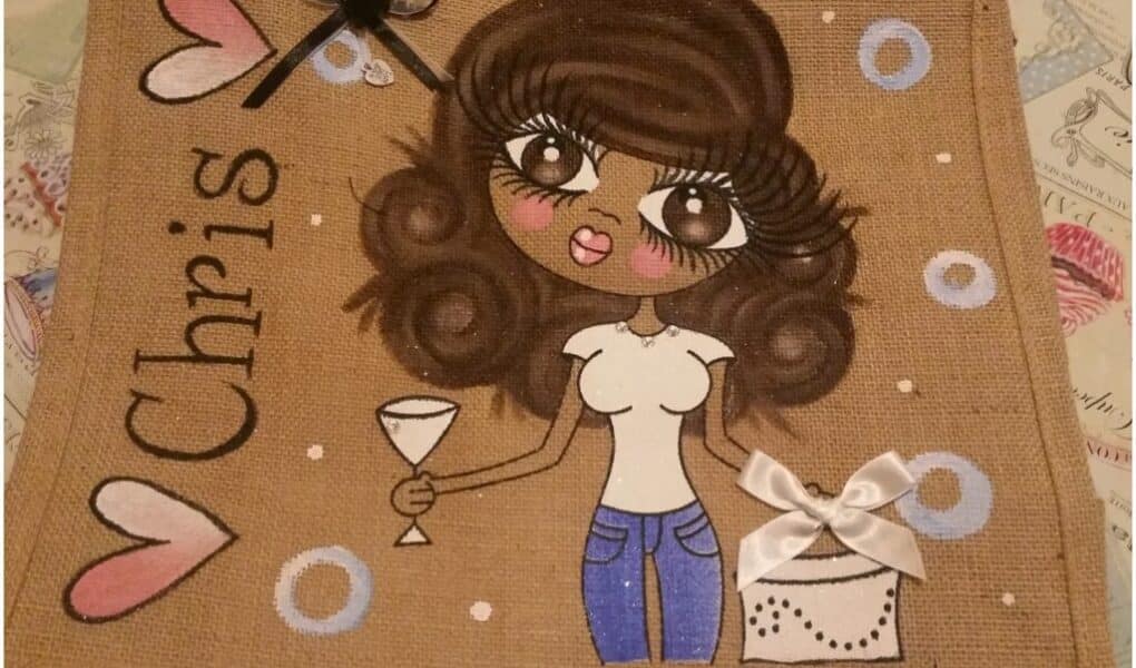 A hessian pouch features a cartoon woman with big wavy brown hair, large eyes, pink lips, a white shirt, and blue jeans. She holds a cocktail glass and stands near a white handbag with a bow. “Chris” and heart shapes decorate the left side.
