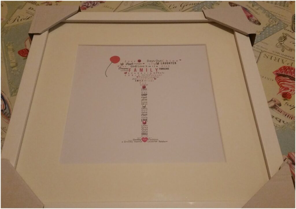 A framed print with a word art tree featuring the word “Family” and phrases about love and laughter, topped with a red balloon. The frame is off-white with cardboard corner protectors, placed on a patterned background with vintage travel and food designs.