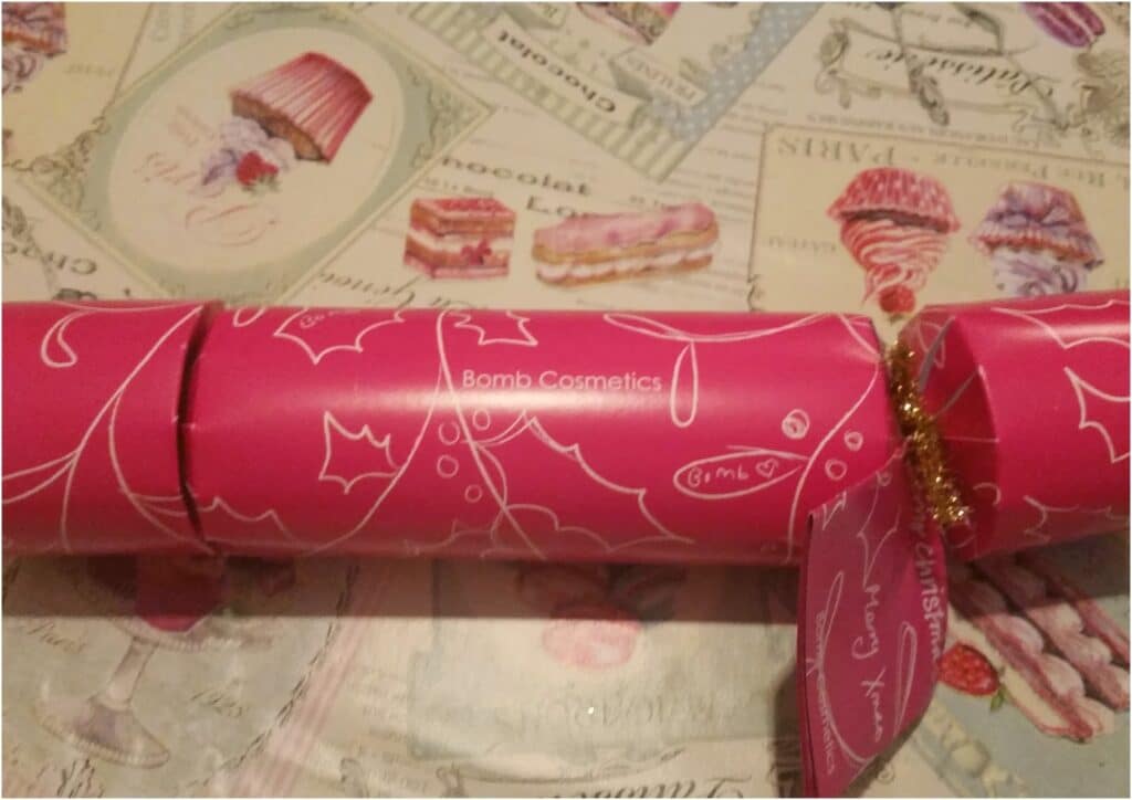 A pink Christmas cracker with white holly designs and "Bomb Cosmetics" printed on it rests on a tablecloth decorated with pastel images and words of cakes, pastries, and desserts. A shiny gold string ties one end of the cracker.