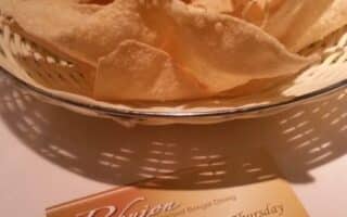 A basket of crispy poppadoms sits on a white tablecloth. In front is a restaurant card for Bhujon, advertising a 4-course meal deal for £11.90 per person, listing meal components and contact information on a gold background.