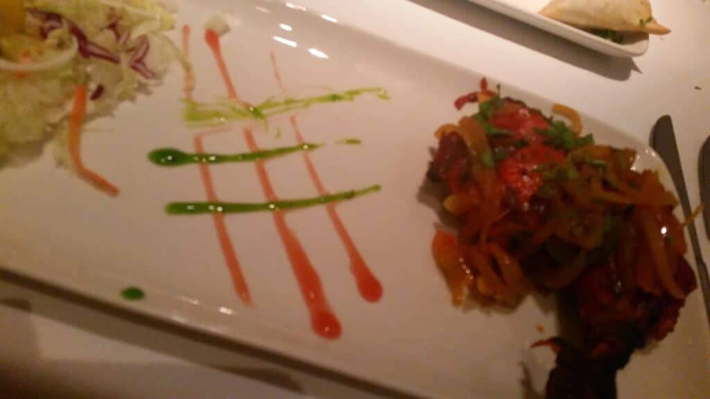A rectangular white plate with shredded cabbage salad on the left, colourful sauce lines—two green and two red—painted diagonally in the centre, and a portion of cooked meat topped with sautéed vegetables on the right. A fork and knife are partly visible.