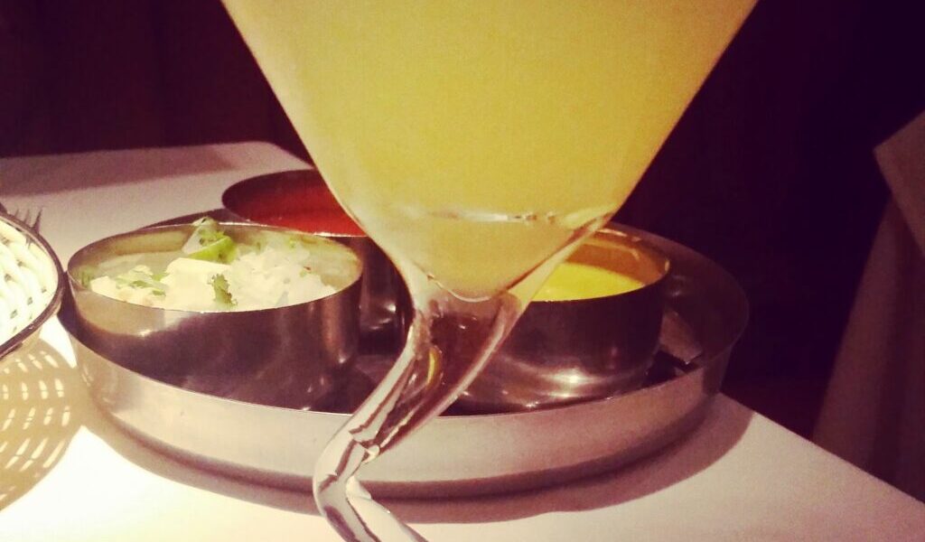 A yellow cocktail served in a martini glass with a twisted stem, set on a white tablecloth. In the background, there are metal bowls containing various condiments, slightly out of focus. The lighting is dim and warm.