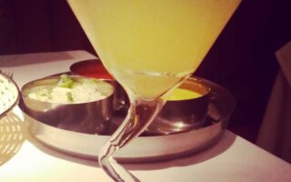 A yellow cocktail served in a martini glass with a twisted stem, set on a white tablecloth. In the background, there are metal bowls containing various condiments, slightly out of focus. The lighting is dim and warm.