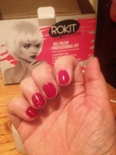 A hand with short, neatly manicured nails painted glossy pink is shown in the foreground. In the background, there is a box labelled "ROKIT Gel Polish Professional Kit" with an image of a woman with a platinum blonde bob haircut.