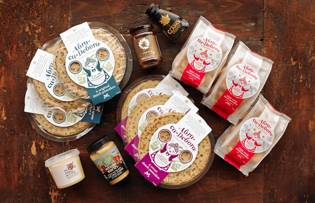 A top-down view of a wooden table displaying stacks of crêpes in round plastic packaging, wrapped galettes, jars of spreads and honey, and bottled syrup. The packaging features whimsical illustrations of women and bold French text.