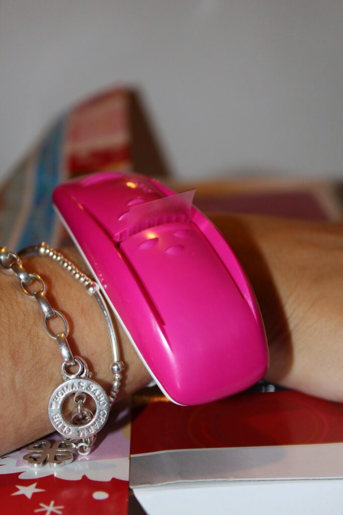 A close-up of a person's wrist wearing silver bracelets and a pink plastic device that dispenses clear tape, with a small piece of tape protruding from its slot. The background is blurred and colourful.