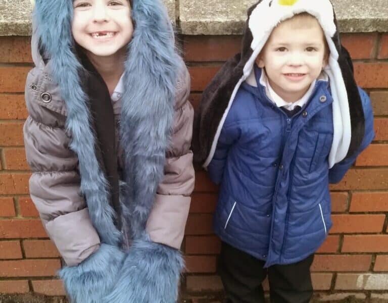 Two young children stand outside against a red brick wall. The child on the left wears a blue fuzzy hat with monster ears and matching paw mittens. The child on the right wears a blue coat and a penguin hat with a yellow beak. Both are smiling.