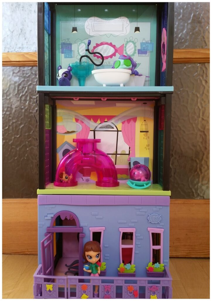A colourful, three-storey toy dolls' house is shown. The top floor has a bathtub and shelves, the middle has a pink slide and hamster ball, and the bottom floor has a balcony with a small doll figure standing on it. The exterior is purple with pink windows and flowers.
