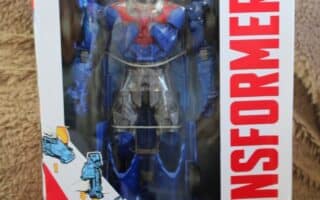 A boxed blue and red Optimus Prime Transformers action figure is displayed upright. The white packaging has a red “TRANSFORMERS” logo on the right, illustrated transformation steps on the left, and the Transformers logo with “Optimus Prime” at the bottom.