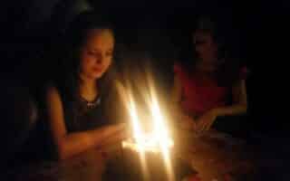 Two young girls sit at a table in a dimly lit room. One girl, with her hands folded, looks down at a lit birthday cake with glowing candles, whilst the other girl, in a red dress, smiles and watches from the background.