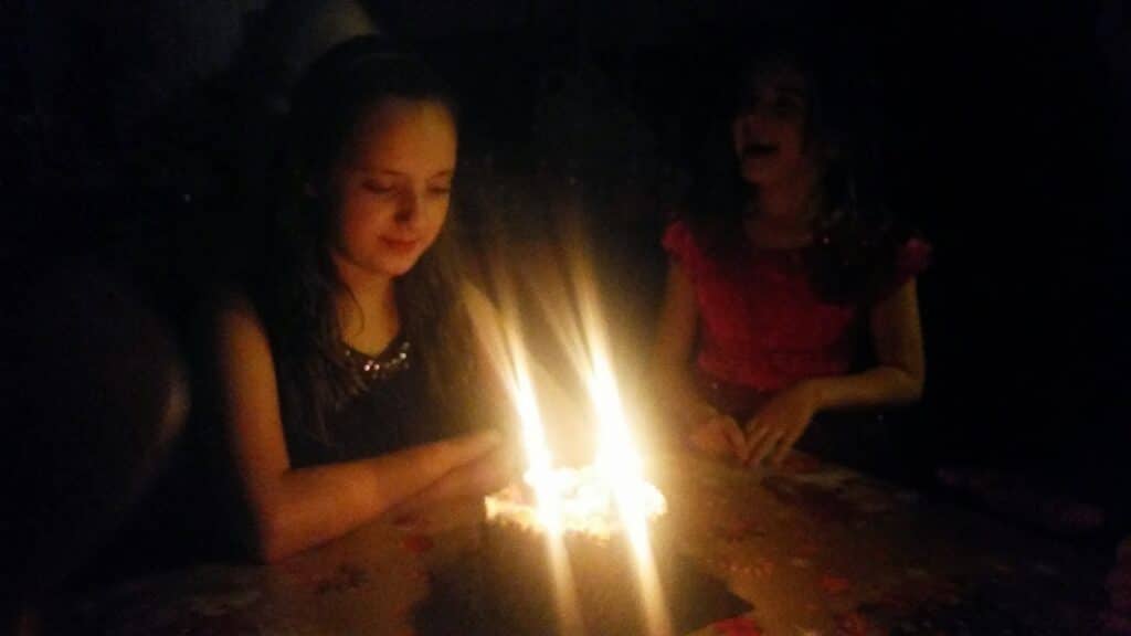 Two young girls sit at a table in a dimly lit room. One girl, with her hands folded, looks down at a lit birthday cake with glowing candles, whilst the other girl, in a red dress, smiles and watches from the background.