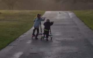 Two children ride scooters on a paved path through a park at sunset. The path is bordered by grass and leafless trees, with soft golden sunlight illuminating the background. The children appear to be playfully interacting as they move forward.