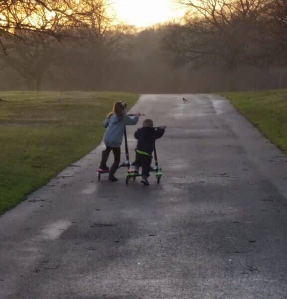 Two children ride scooters on a paved path through a park at sunset. The path is bordered by grass and leafless trees, with soft golden sunlight illuminating the background. The children appear to be playfully interacting as they move forward.