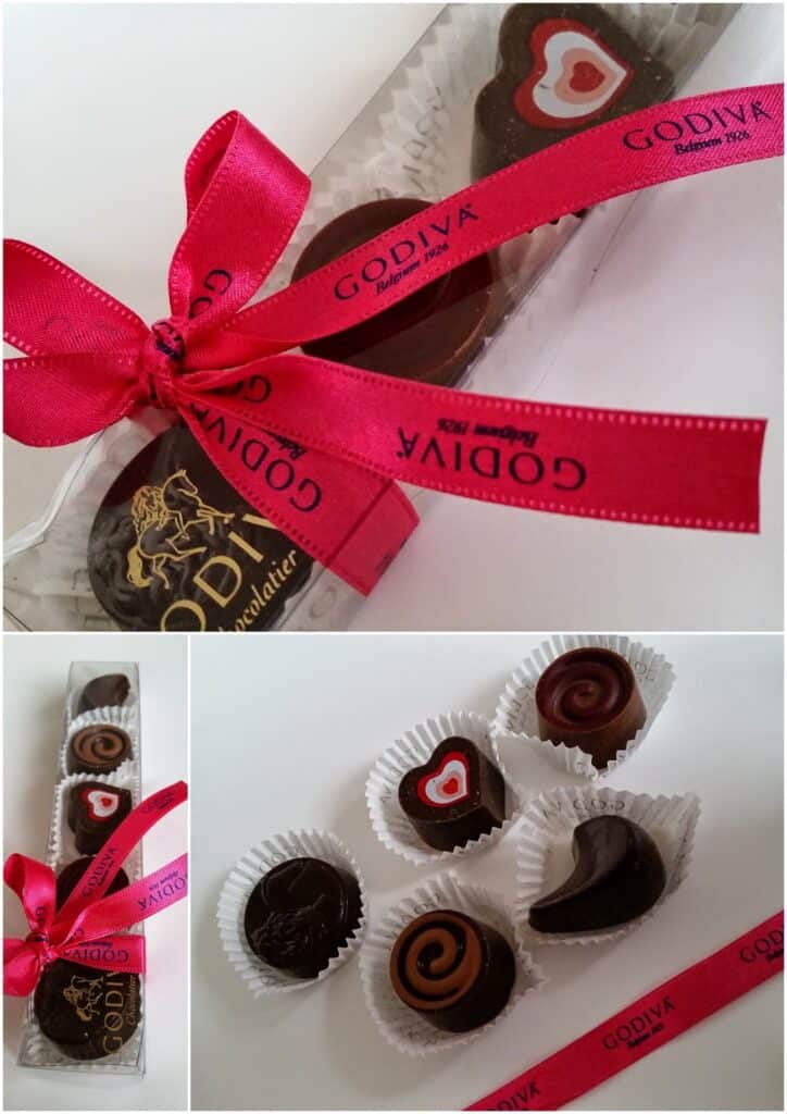 A clear box of assorted Godiva chocolates is tied with a bright pink ribbon and bow. Some chocolates have heart designs. An open view shows five chocolates with varied shapes and decorations, placed in white paper cases.