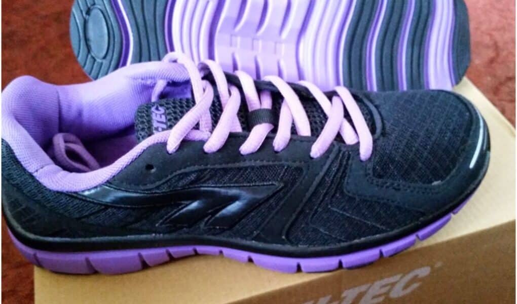 A pair of black trainers with purple soles, purple laces, and purple lining are placed on a cardboard HI-TEC shoe box. One shoe stands upright while the other is laid on its side, showing the sole. The background is a reddish surface.