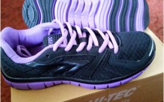A pair of black trainers with purple soles, purple laces, and purple lining are placed on a cardboard HI-TEC shoe box. One shoe stands upright while the other is laid on its side, showing the sole. The background is a reddish surface.