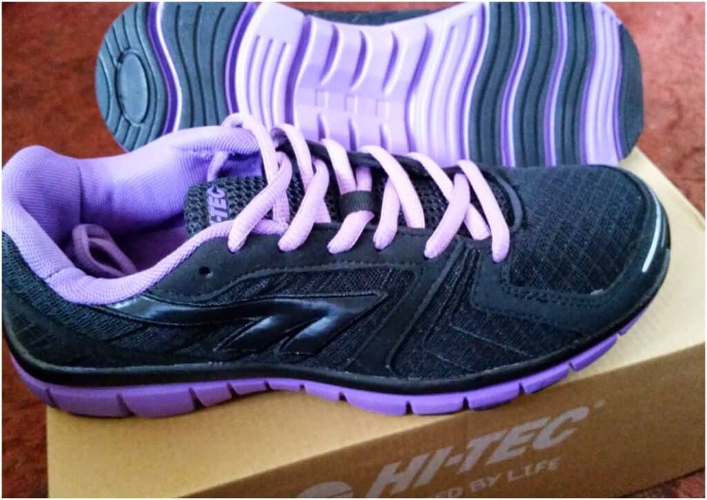A pair of black and purple athletic trainers with purple laces and soles, placed on a tan HI-TEC shoe box. One shoe stands upright while the other is propped behind it, showing the textured sole. The shoes are set on a reddish-brown surface.