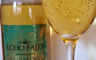 A bottle of Echo Falls Alcohol Free Sparkling Infusion with a gold and teal label stands next to a wine glass filled with bubbly, golden liquid. The label reads “ZERO Alcohol” and “Sparkling Infusion”.