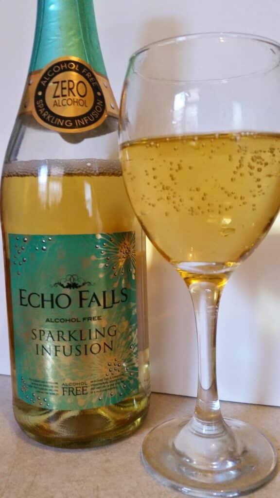 A bottle of Echo Falls Alcohol-Free Sparkling Infusion sits next to a wine glass filled with its golden, bubbly liquid. The bottle has a green and gold label, with "Zero Alcohol" and decorative sparkling patterns visible.