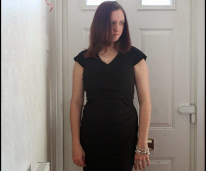 A woman with straight brown hair stands indoors in front of a white door, wearing a fitted black dress and silver bracelets. She looks slightly to her left with a neutral expression. Red ribbon decoration hangs in the top left corner. Light enters from the door’s window.