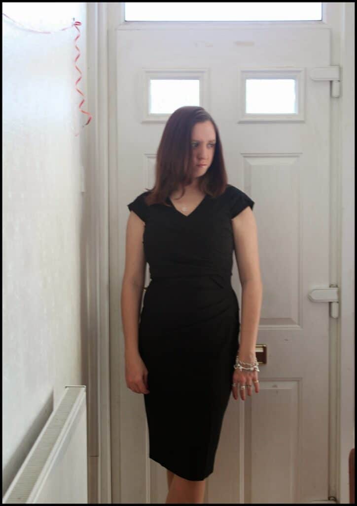A woman with straight brown hair wearing a fitted black dress stands indoors in front of a white door. She has a neutral expression, her left arm relaxed by her side, and she wears bracelets on her left wrist. Red ribbon decorates the top left corner.