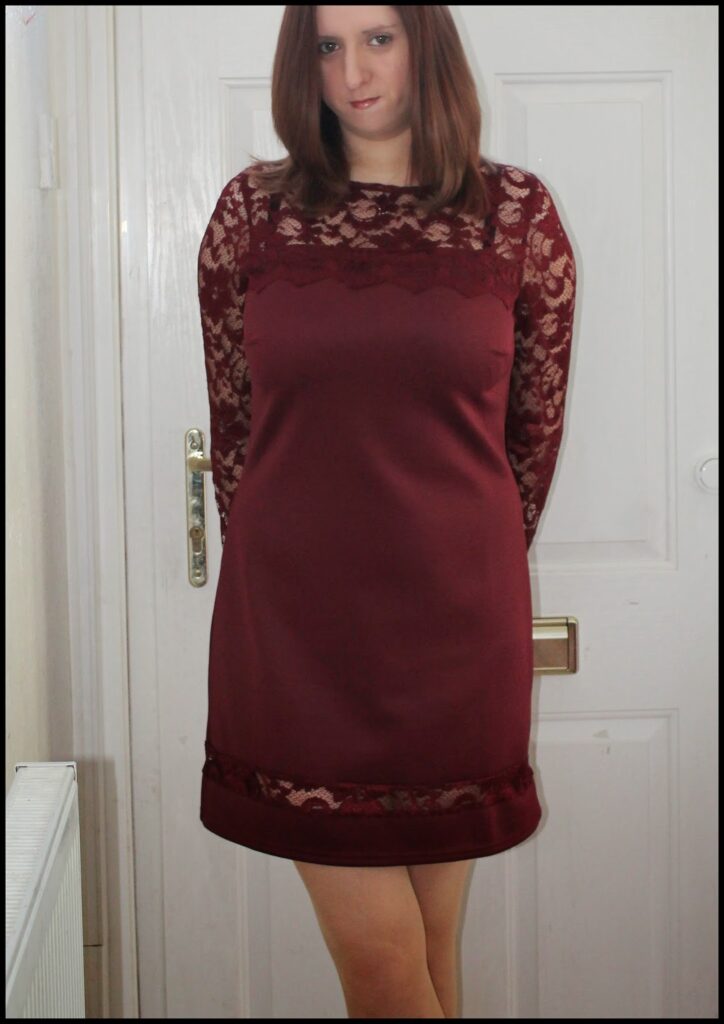 A woman with straight brown hair stands indoors in front of a white door. She is wearing a knee-length, long-sleeved burgundy dress with lace detailing on the sleeves and upper chest. Her arms are behind her back, and she is looking slightly at the camera.