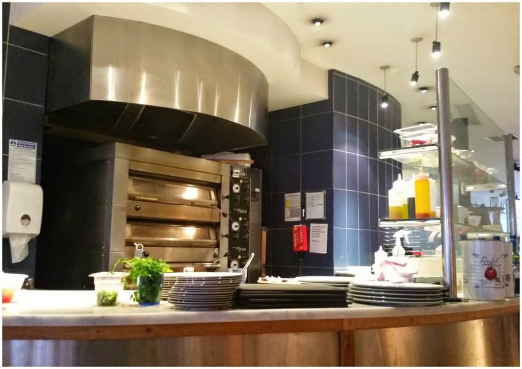A commercial kitchen with a large stainless steel pizza oven, blue tiled walls, stacked white plates on a counter, containers of sauces, herbs, and condiments, and bright overhead lighting creating a clean, organised workspace.