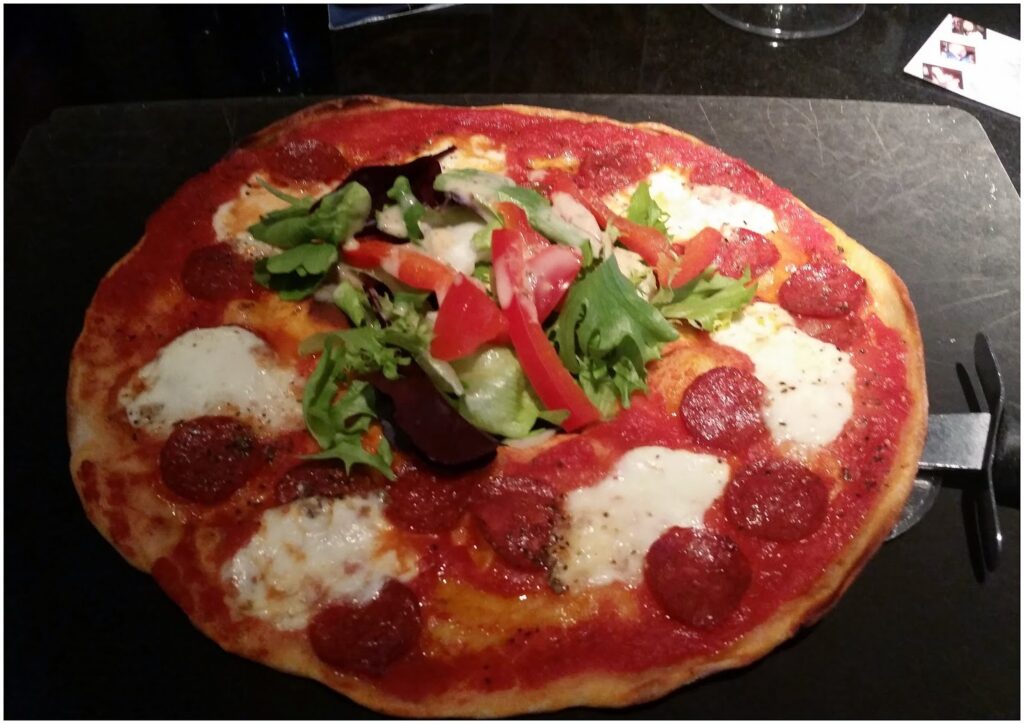 A round pizza topped with pepperoni slices, melted mozzarella cheese, and a generous pile of fresh mixed leaves, tomato, and red pepper in the centre, served on a dark surface with part of a pizza cutter visible on the right side.