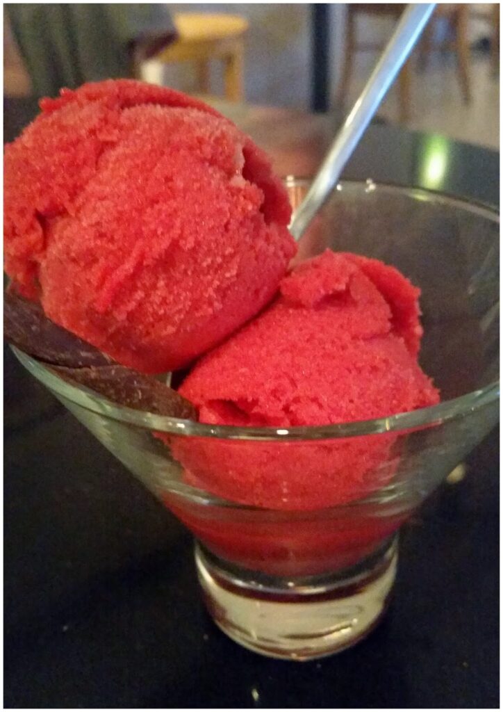 A glass bowl containing two scoops of bright red sorbet sits on a dark surface. A metal spoon is inserted into one scoop. The background is blurred, showing brown and tan chairs and tables, suggesting a casual indoor setting.