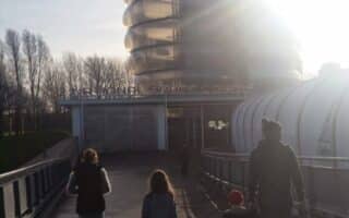 Four people, two adults and two children, walk on a sunlit path towards the National Space Centre in Leicester, UK. The building’s distinctive, rounded Rocket Tower structure rises ahead, partially obscured by bright sunlight and long shadows.