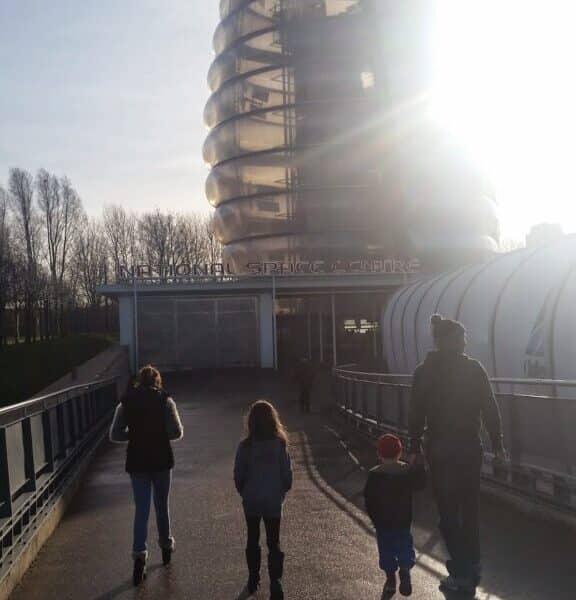 Four people, two adults and two children, walk on a sunlit path towards the National Space Centre in Leicester, UK. The building’s distinctive, rounded Rocket Tower structure rises ahead, partially obscured by bright sunlight and long shadows.
