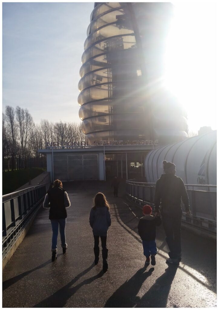 Four people, including two children, walk towards a futuristic glass building with a curved design in bright sunlight. Shadows stretch ahead of the group. The building is labelled “National Space Centre.” Leafless trees are visible in the background.