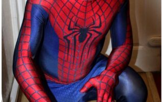 A person dressed in a full Spider-Man costume crouches indoors near a white door, with one hand extended forwards and the other bent, imitating Spider-Man's iconic web-shooting pose. The suit is red and blue with a web pattern and a large black spider emblem on the chest.