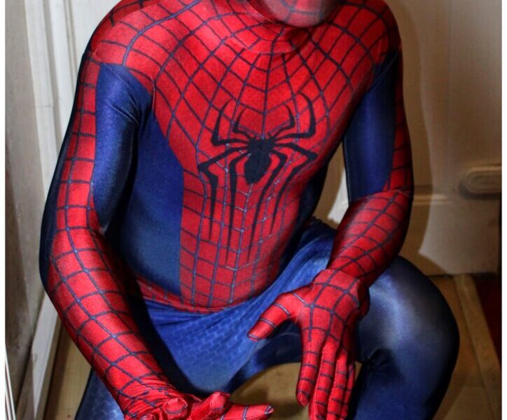 A person dressed in a full Spider-Man costume crouches indoors near a white door, with one hand extended forwards and the other bent, imitating Spider-Man's iconic web-shooting pose. The suit is red and blue with a web pattern and a large black spider emblem on the chest.