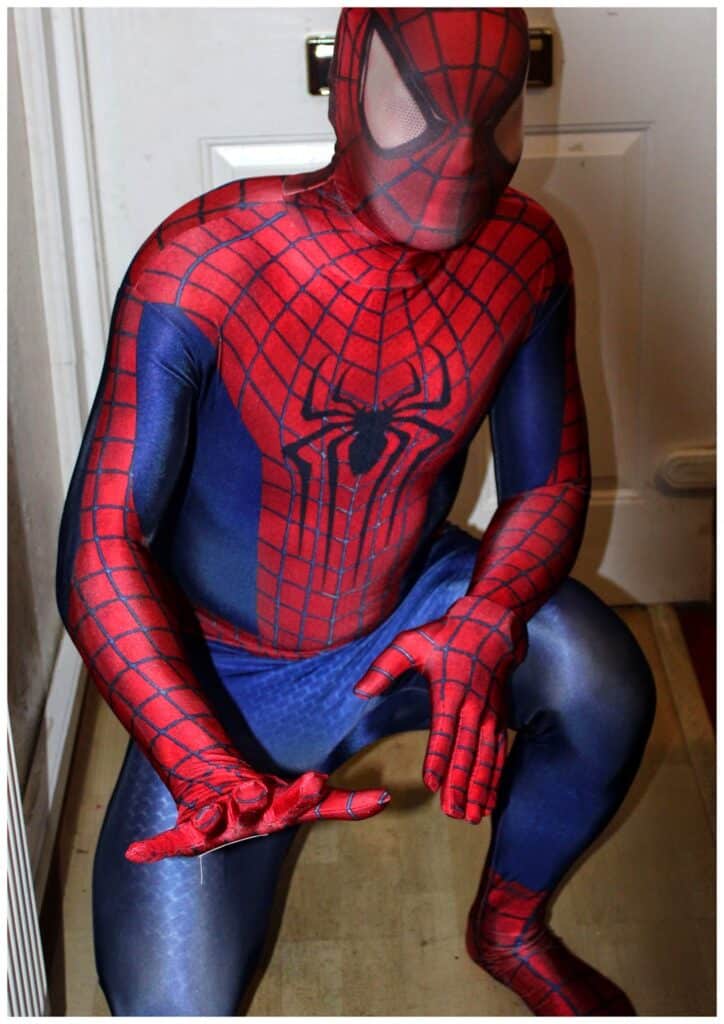 A person wearing a full-body Spider-Man costume, featuring a red and blue suit with a web pattern and spider emblem, crouches in front of a white door, mimicking Spider-Man's iconic web-shooting pose with one hand extended.