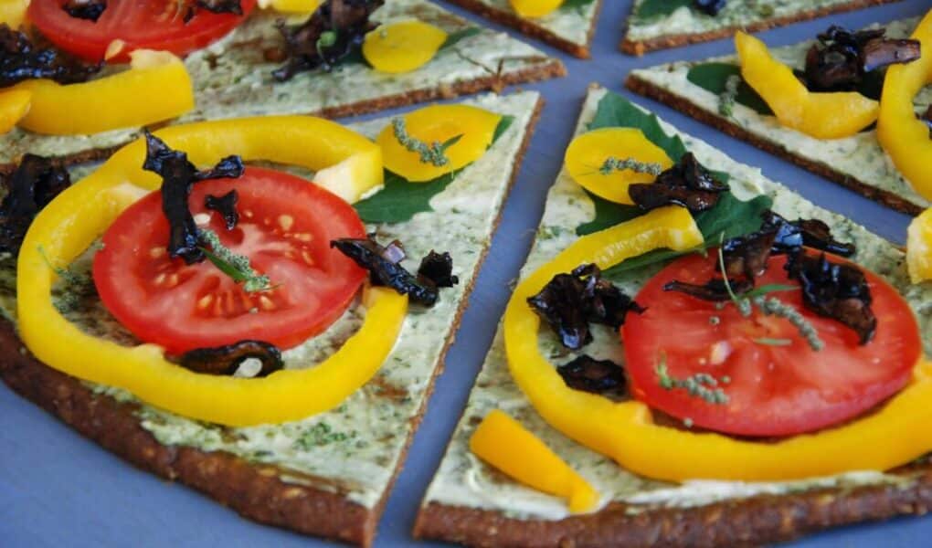 A close-up of sliced pizza topped with fresh tomato slices, yellow pepper rings, dark mushrooms, and herbs on a thin, greenish crust, all arranged on a blue surface. The pizza appears colourful, fresh, and vegetable-rich.