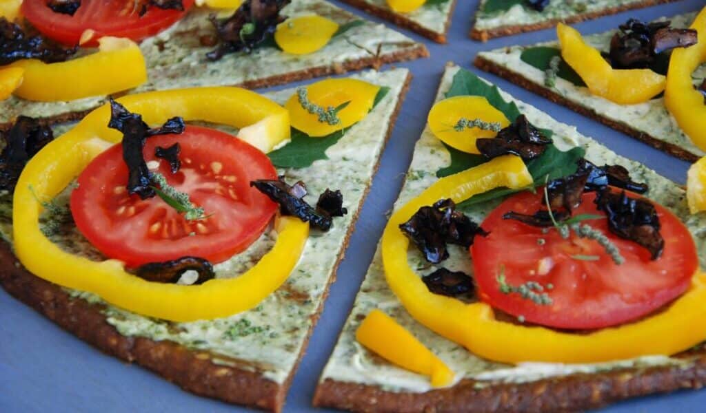 A close-up of sliced pizza topped with fresh tomato slices, yellow pepper rings, dark mushrooms, and herbs on a thin, greenish crust, all arranged on a blue surface. The pizza appears colourful, fresh, and vegetable-rich.
