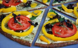 A close-up of sliced pizza topped with fresh tomato slices, yellow pepper rings, dark mushrooms, and herbs on a thin, greenish crust, all arranged on a blue surface. The pizza appears colourful, fresh, and vegetable-rich.