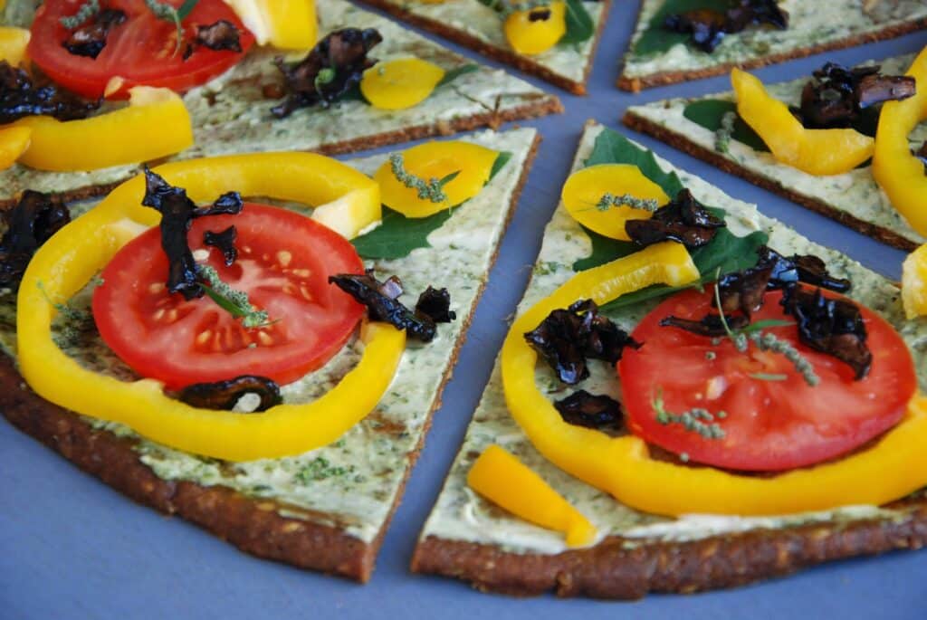 A close-up of sliced vegetable pizza topped with yellow pepper rings, tomato slices, dark mushrooms, leafy greens, and herbs on a thin, crispy base. The colourful toppings are fresh and vibrant, arranged neatly on each slice.