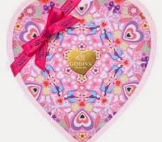 Heart-shaped box with a colourful, floral kaleidoscope pattern in pink, purple, and blue tones. A gold Godiva logo is at the centre, and a pink ribbon with a bow is tied on the upper left corner for decoration.
