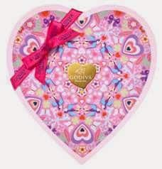 Heart-shaped box with a colourful, floral kaleidoscope pattern in pink, purple, and blue tones. A gold Godiva logo is at the centre, and a pink ribbon with a bow is tied on the upper left corner for decoration.