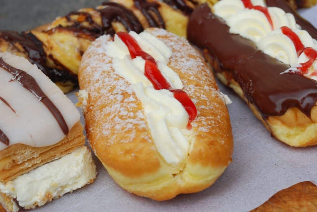 Three assorted cream-filled pastries are shown: one topped with white icing, one dusted with icing sugar, filled with cream and a red jam stripe, and one covered in chocolate icing with white and red cream decoration, all on baking parchment.